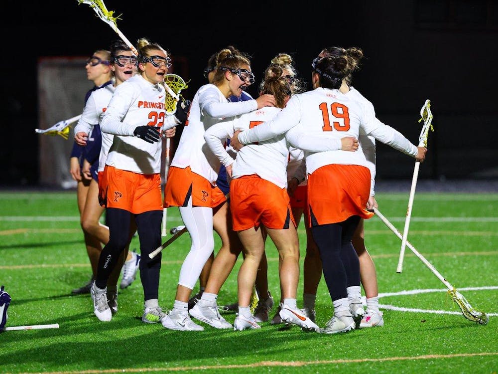 Huddle of Princeton lacrosse players with white jerseys and orange shorts gather on the green field. The girls are hold lacrosse sticks. 