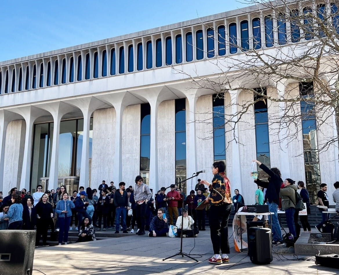 A crowd stands in front of a white building with columns facing a woman standing at a microphone
