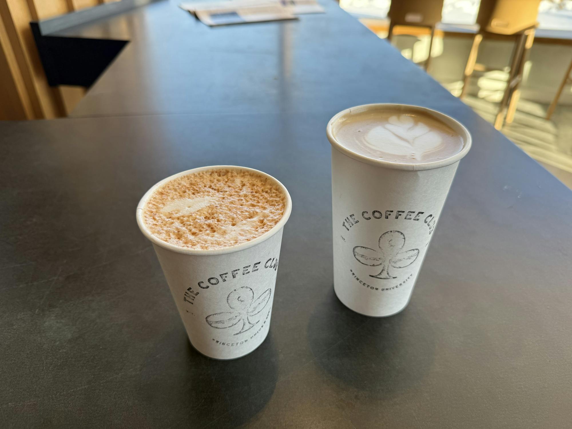 Two cardboard coffee cups, one filled with a foamy drink and the other with a latte swirl, sits on the table.