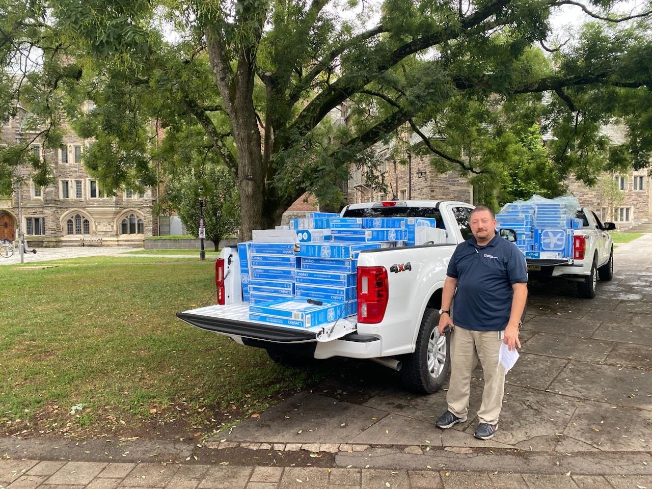 Man in navy shirt stands in front of two white trucks filled with fans in blue boxes.