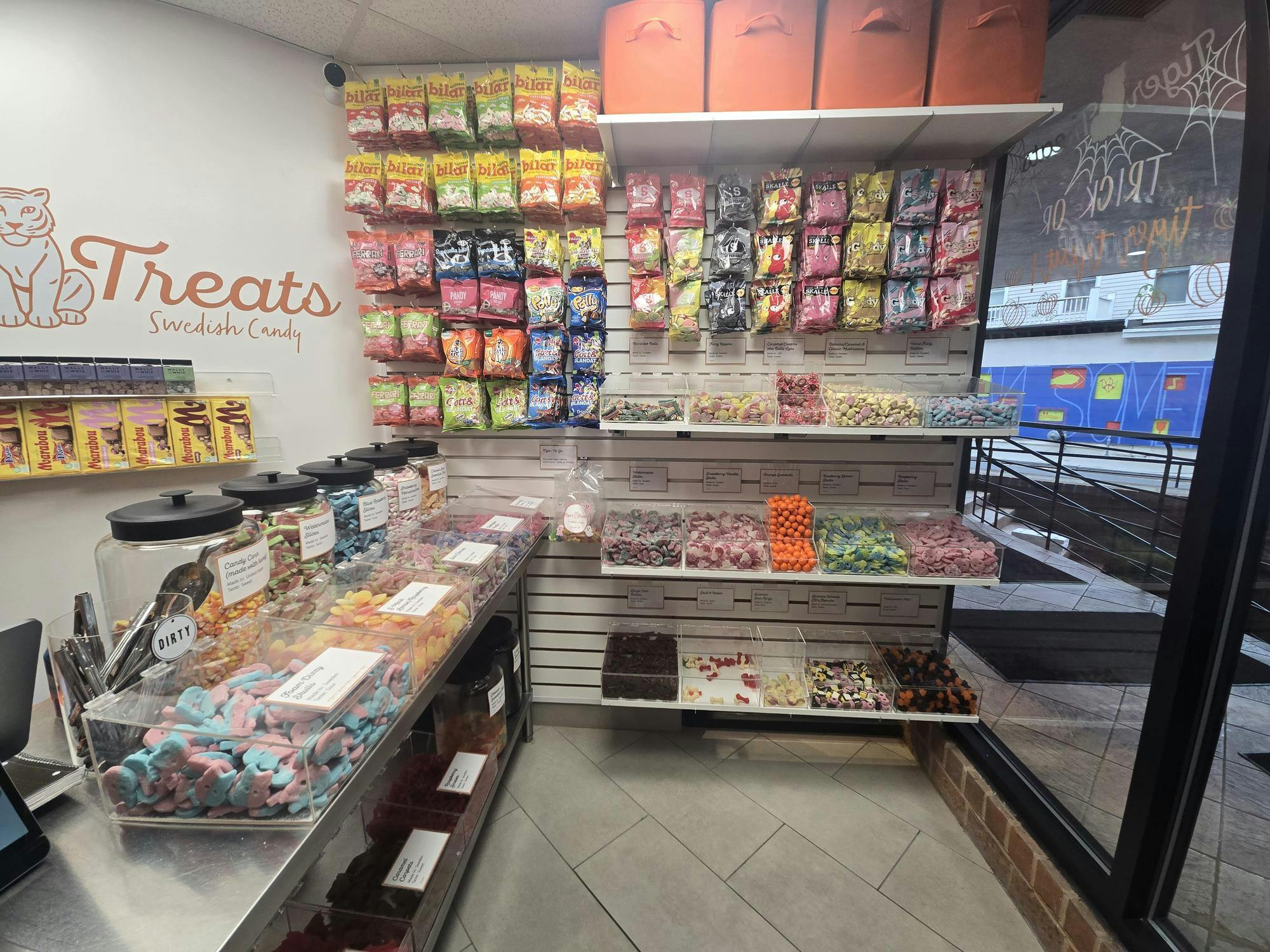 Multicolored bags of candy are hanging on a white wall. There is a Tiger Treats sign in orange script font on the adjacent white wall.