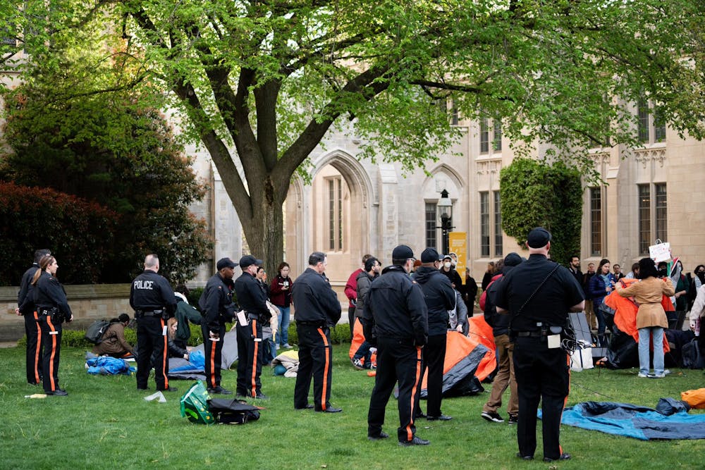 A line of people in black shirts and black pants with an orange stripe down the leg stand in a line. They face a group of people holding tents and signs. 