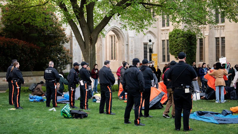 A line of people in black shirts and black pants with an orange stripe down the leg stand in a line. They face a group of people holding tents and signs. 