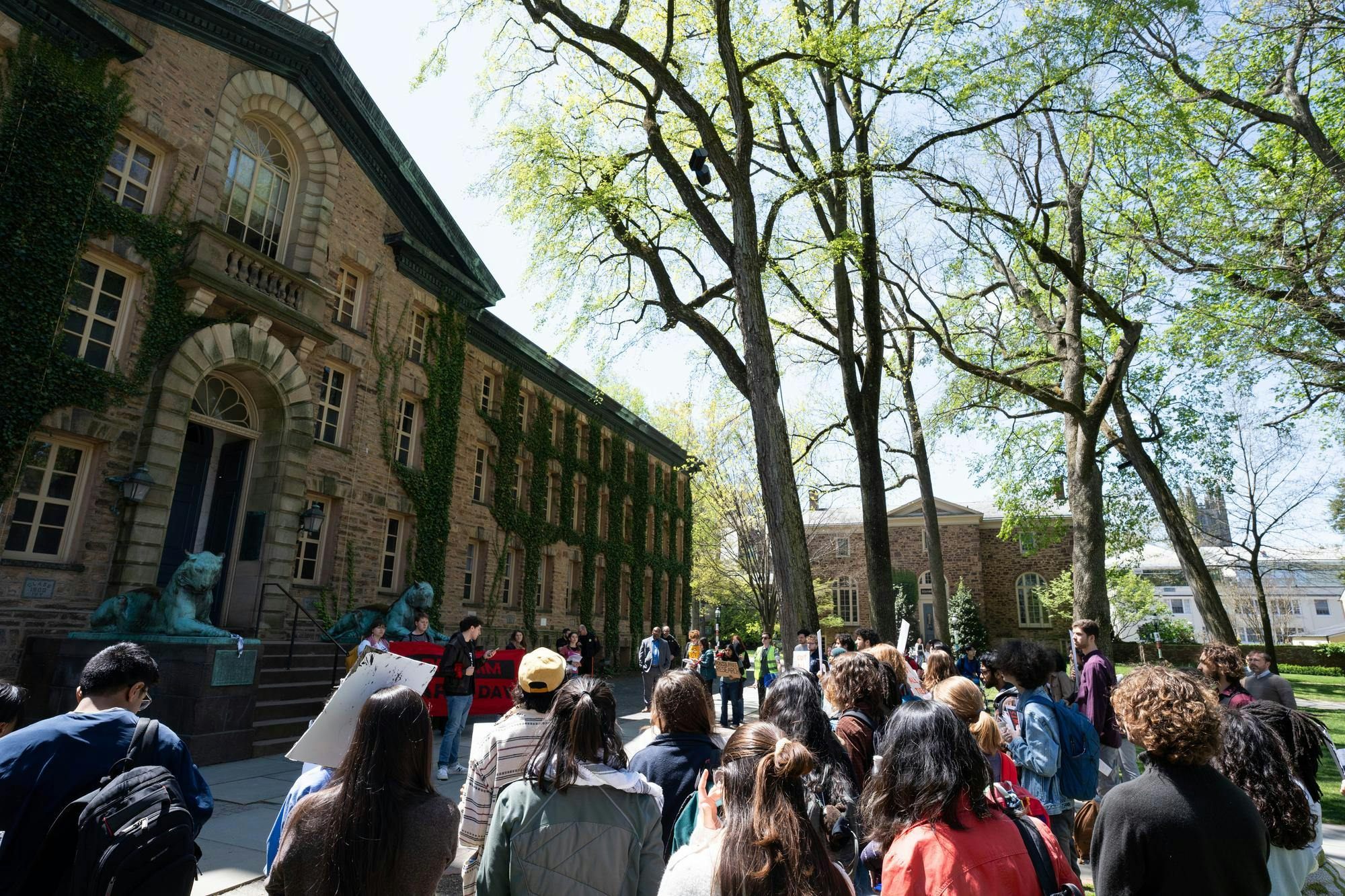 In the background on the right, a building composed of brown bricks covered with green ivy has a large black door in front. On the right lower third foreground, many protestors gather in front of a man speaking. In the top right, lightly leafed trees sit below a bright sun and pale blue sky.