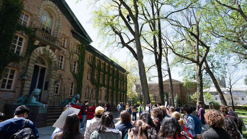 In the background on the right, a building composed of brown bricks covered with green ivy has a large black door in front. On the right lower third foreground, many protestors gather in front of a man speaking. In the top right, lightly leafed trees sit below a bright sun and pale blue sky.