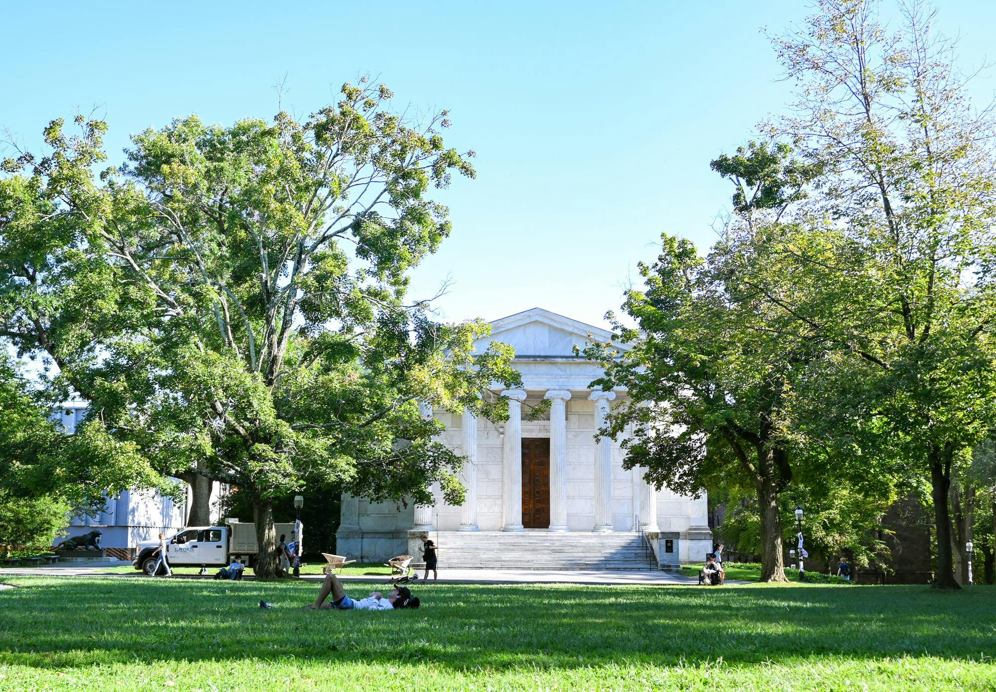 Pictured is a white building with large white columns and steps in front. There are two large trees on both sides of the building and a large area of grass in front of it.
