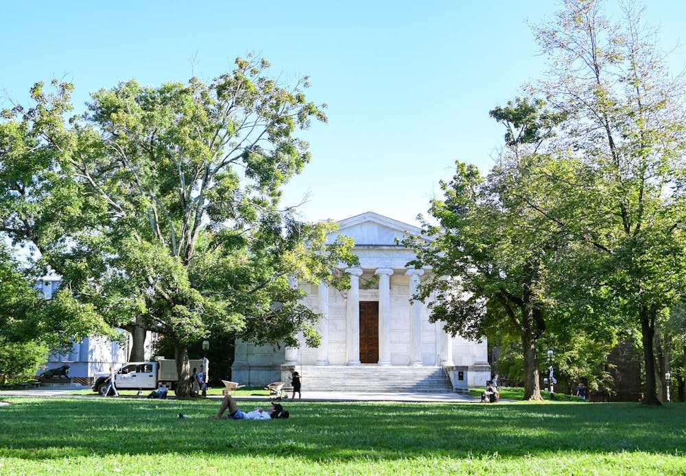 Pictured is a white building with large white columns and steps in front. There are two large trees on both sides of the building and a large area of grass in front of it.