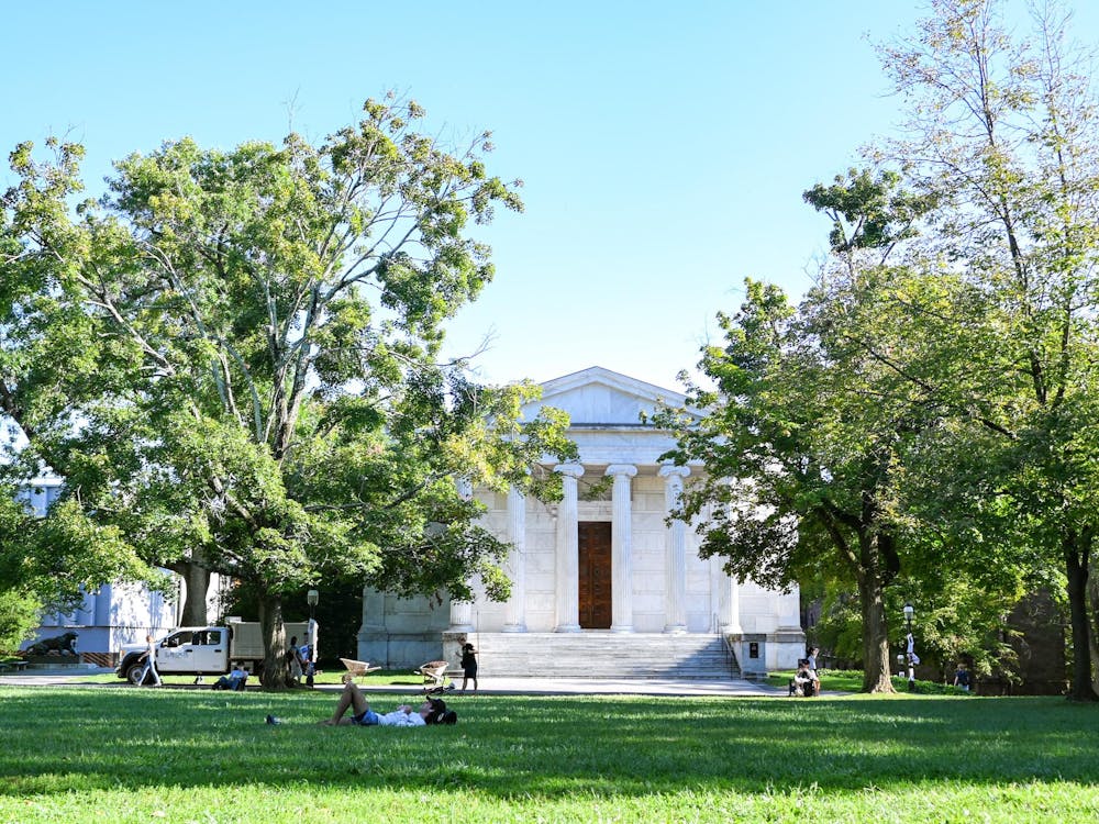 Pictured is a white building with large white columns and steps in front. There are two large trees on both sides of the building and a large area of grass in front of it.