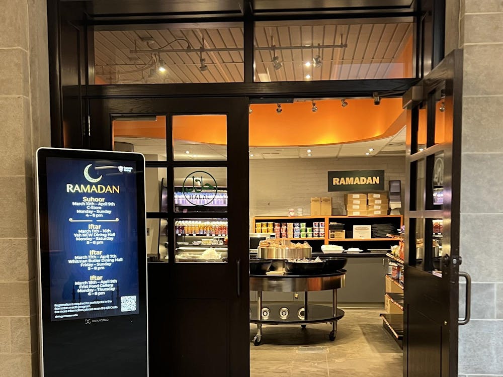 A set of two doors with a green marking in the window that reads “Halal,” and numerous signs in the background and foreground that say “Ramadan.” Past the doors lie a set of tables and shelves with various food items on them.