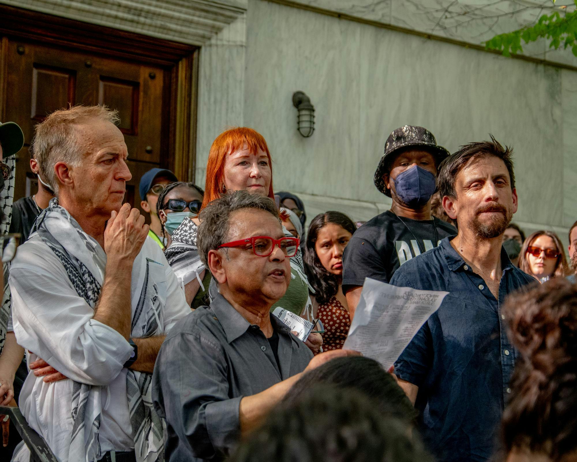 Four people watch a woman read a sheet of paper, surrounded by a crowd of students.