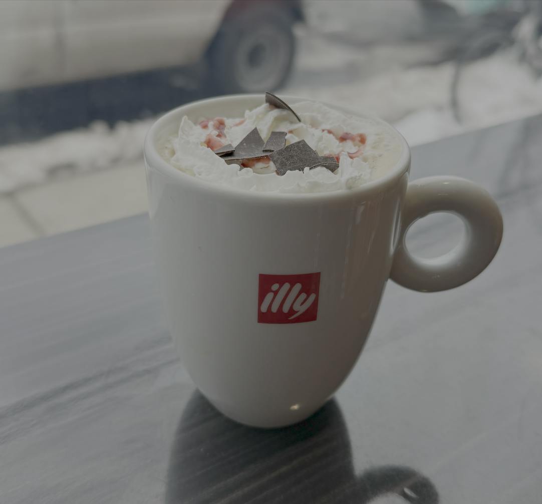 A warm beverage sits in a white mug, topped with whipped cream, chocolate shavings, and heart-shaped red and pink sprinkles. 