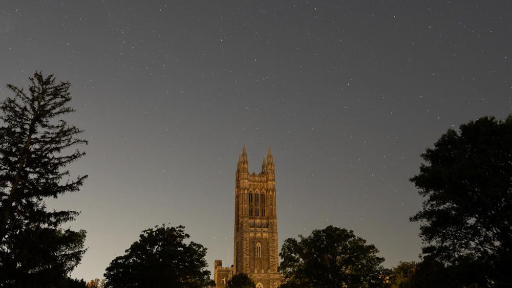 This is a photo taken at night with Cleveland Tower, a tall gothic tower that's part of Princeton's graduate college, in the foreground. Trees surround the base of the tower, and a few stars are visible in the sky. 
