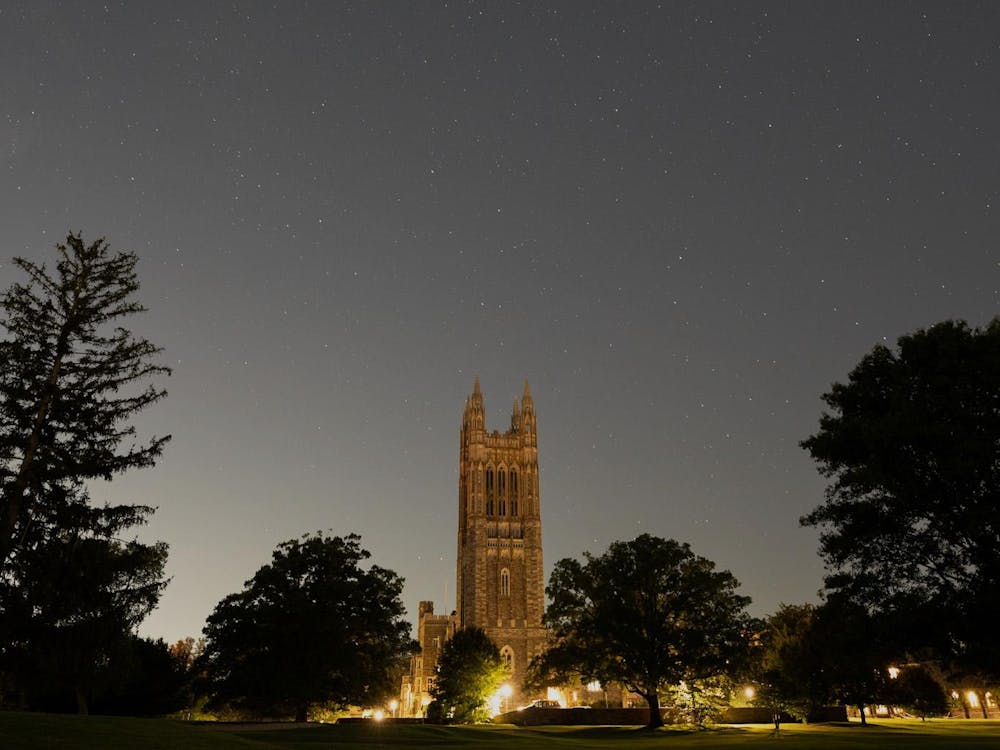 This is a photo taken at night with Cleveland Tower, a tall gothic tower that's part of Princeton's graduate college, in the foreground. Trees surround the base of the tower, and a few stars are visible in the sky. 