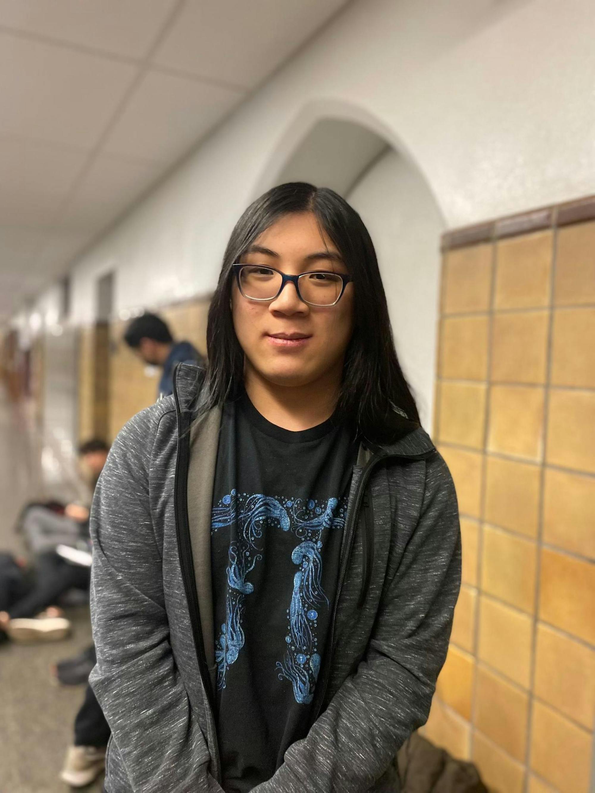 A teenager with glasses and a gray sweatshirt stands in a hallway with brown tiled walls