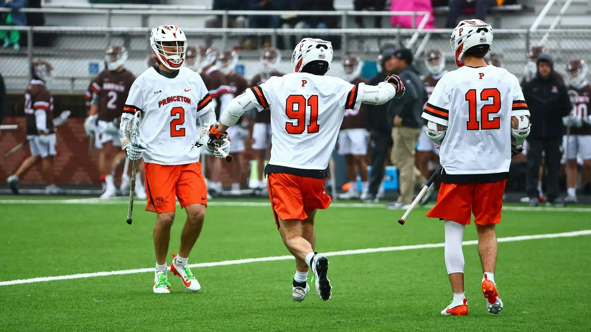 Three men in orange shorts and white jerseys on a grass field holding lacrosse sticks.