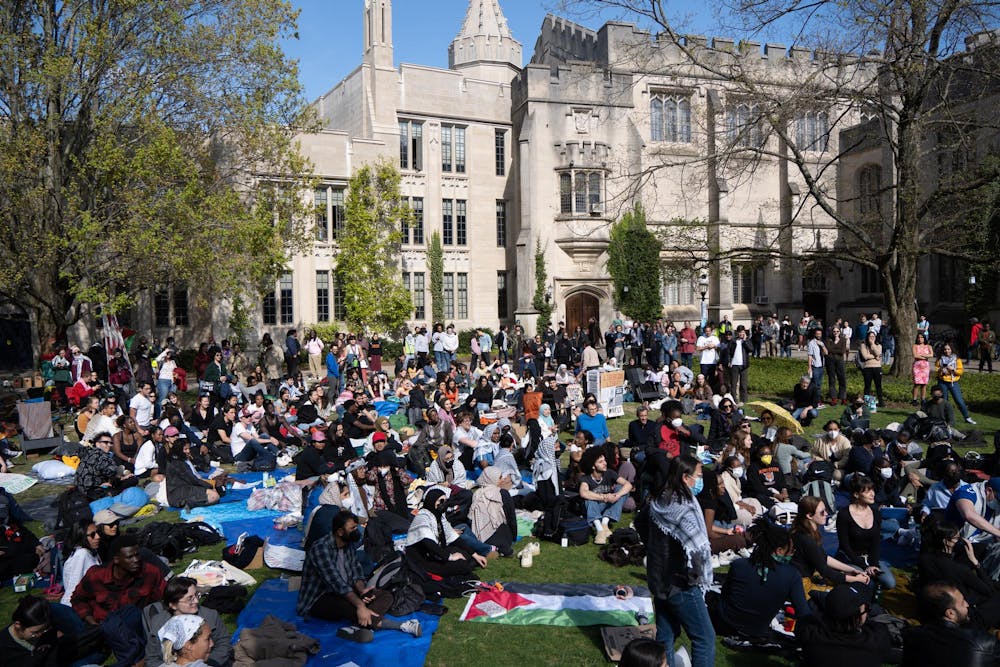 Students sit on lawn with picnic blankets and Palestinian red, green, white, and black flags. They lie in from of a Tudor Gothic building. 