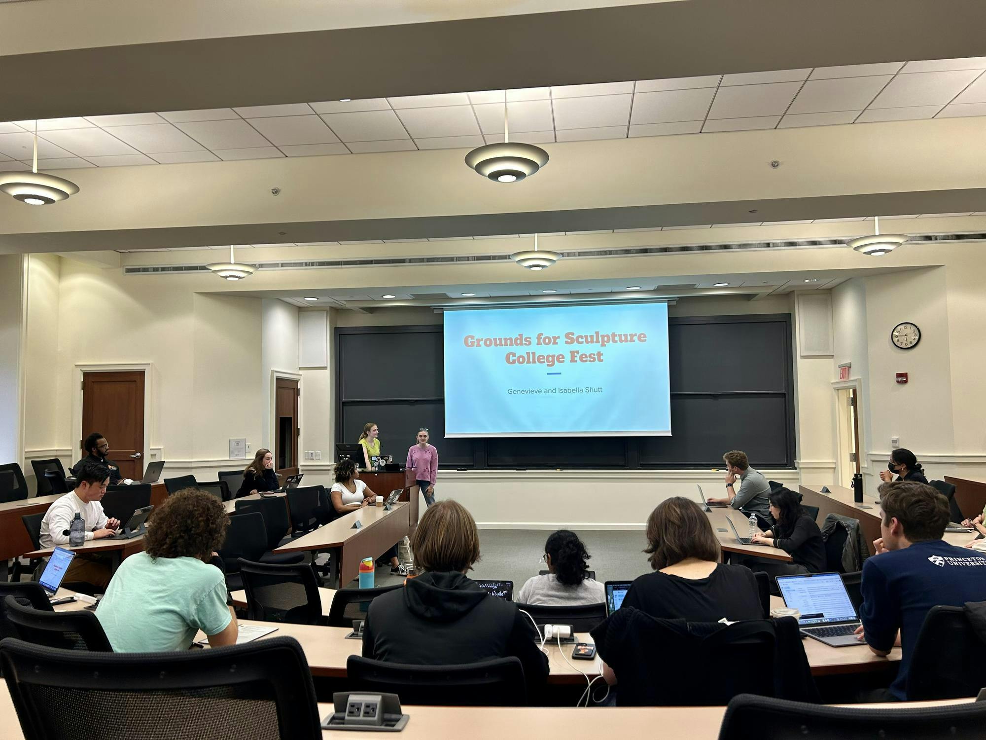 Students sit in a conference room with laptops in front of them with a PowerPoint on the screen saying "Grounds for Sculpture College Fest."