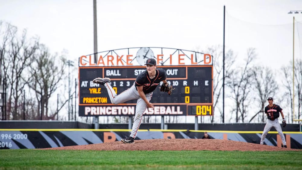 A player with a black jersey stands on a baseball mound on one leg.