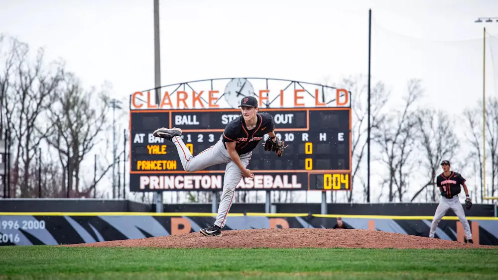 A player with a black jersey stands on a baseball mound on one leg.