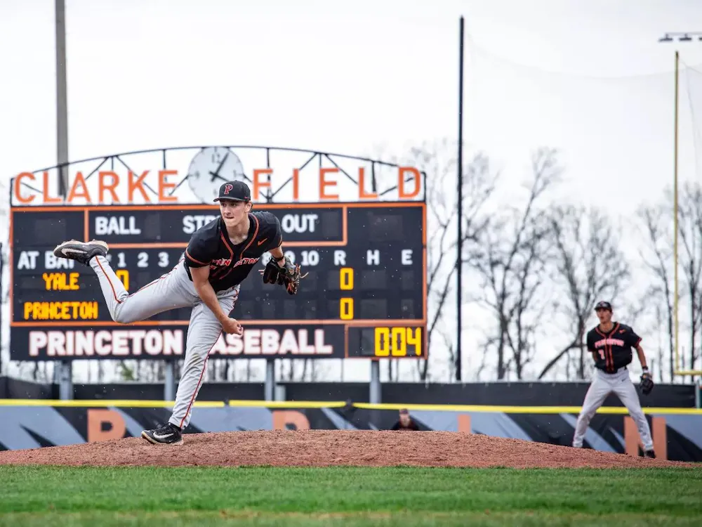 A player with a black jersey stands on a baseball mound on one leg.