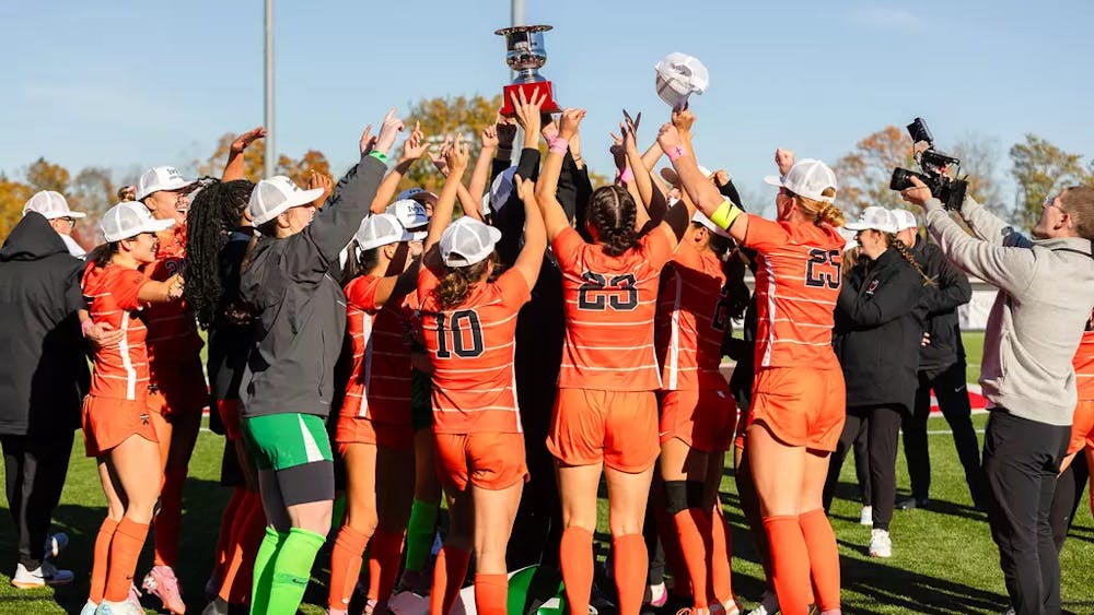 Women’s soccer team in orange jerseys celebrate in a circle and hoist a trophy.