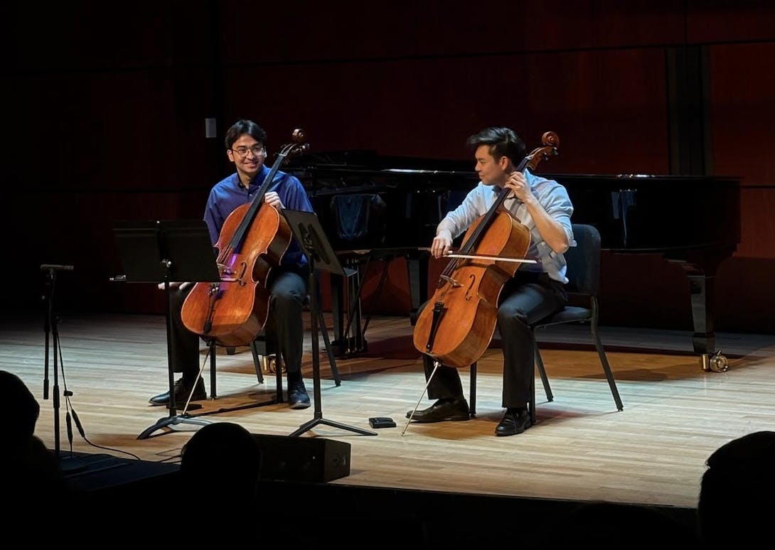 Two young men each play a cello, seated in black chairs on a wooden stage. The man on the right has dark hair, wears a dark blue shirt, and is smiling at the man on the left who is wearing a lighter blue shirt and also has dark hair. 