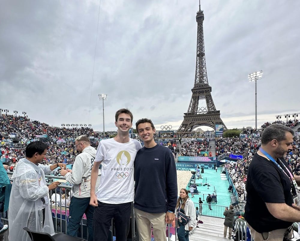 Two people posing inside of a crowded stadium with a crowd and tall metal tower behind.