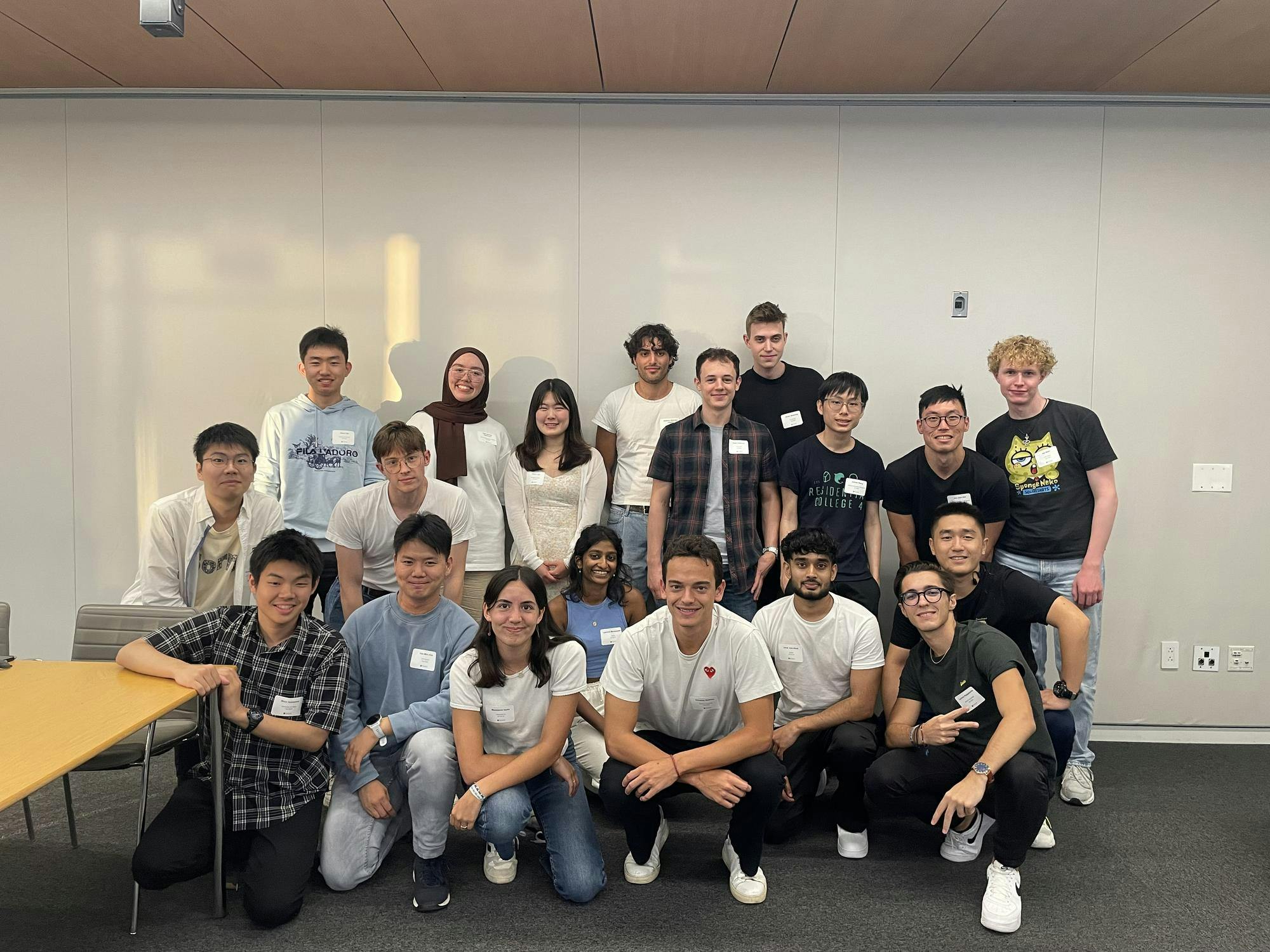 19 students pose in front of a white background. They wear casual clothing. 