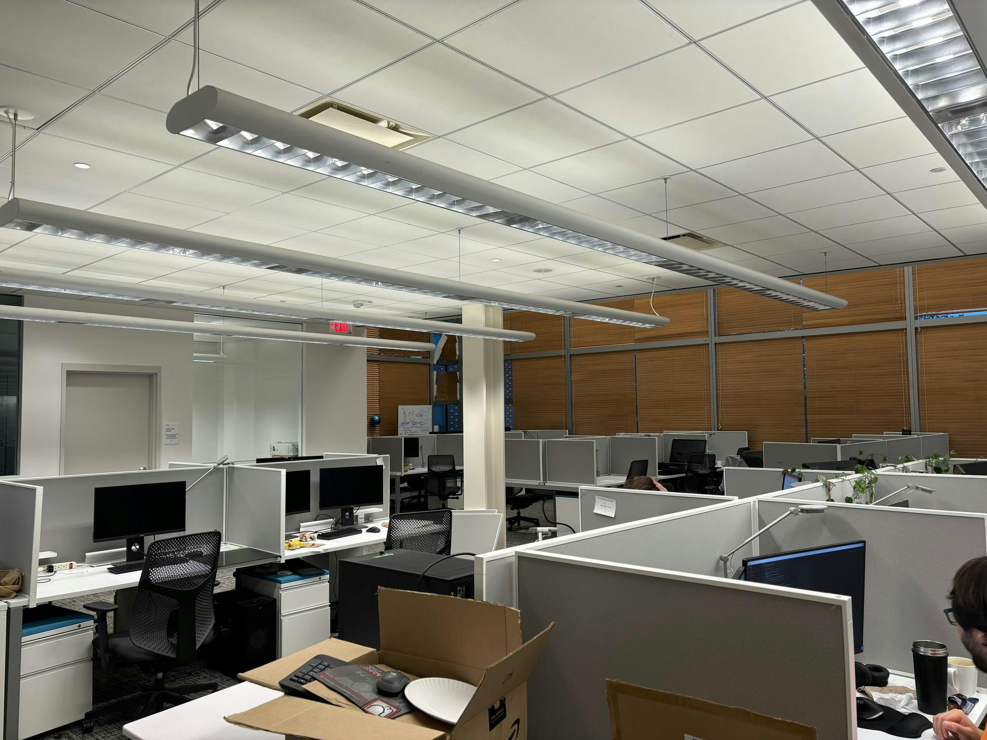 Gray cubicles with computers are lined up in rows in a basement room. 