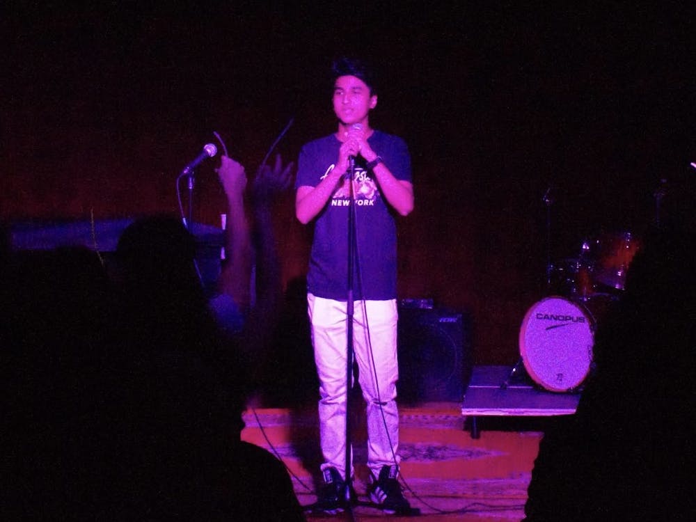 A young man stands at a microphone stand, with his hands wrapped around the microphone, on a dark stage with purple and pink spotlights shining onto him.