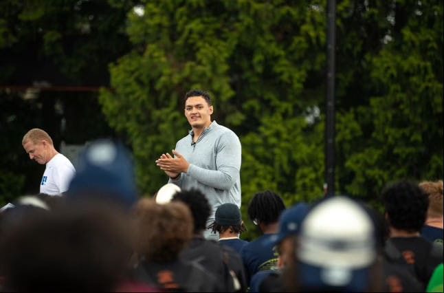 A man with a gray sweater clapping in front of an audience. 