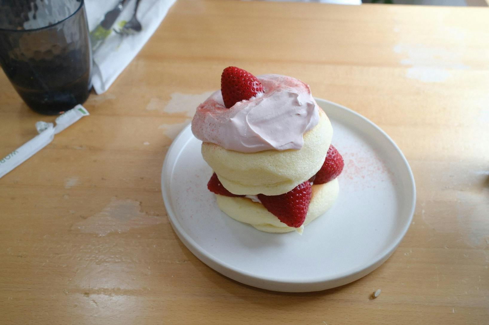 A small white plate has a stack of light yellow fluffy pancakes. There are red strawberries and white cream throughout the stack. The whole plate sits on a light brown table.