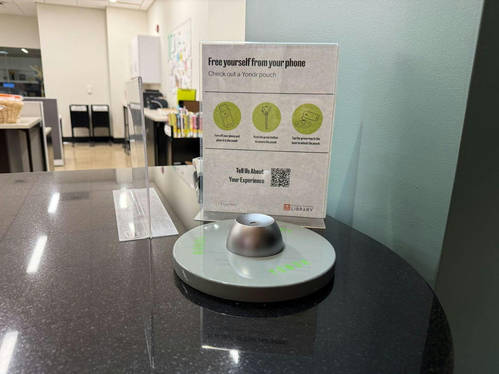 A paper advertises the Yondr phone pouch to students. In bold, it says "Free yourself from your phone." Below it, it says "Check out a Yondr pouch." In front of the paper is a device to unlock pouches.