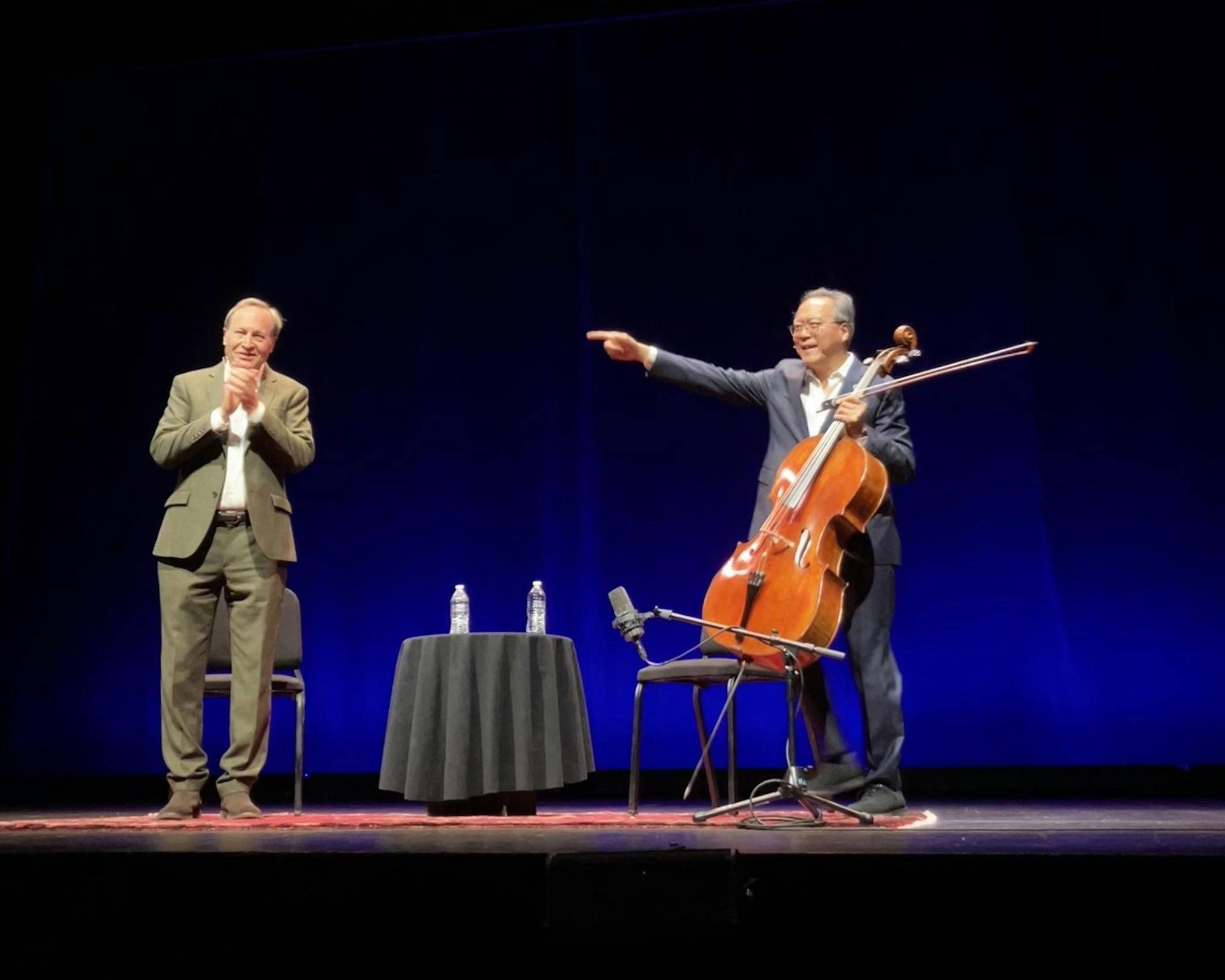 A man on the right holds a cello and a cello bow, while a man on the left is clapping his hands together. 