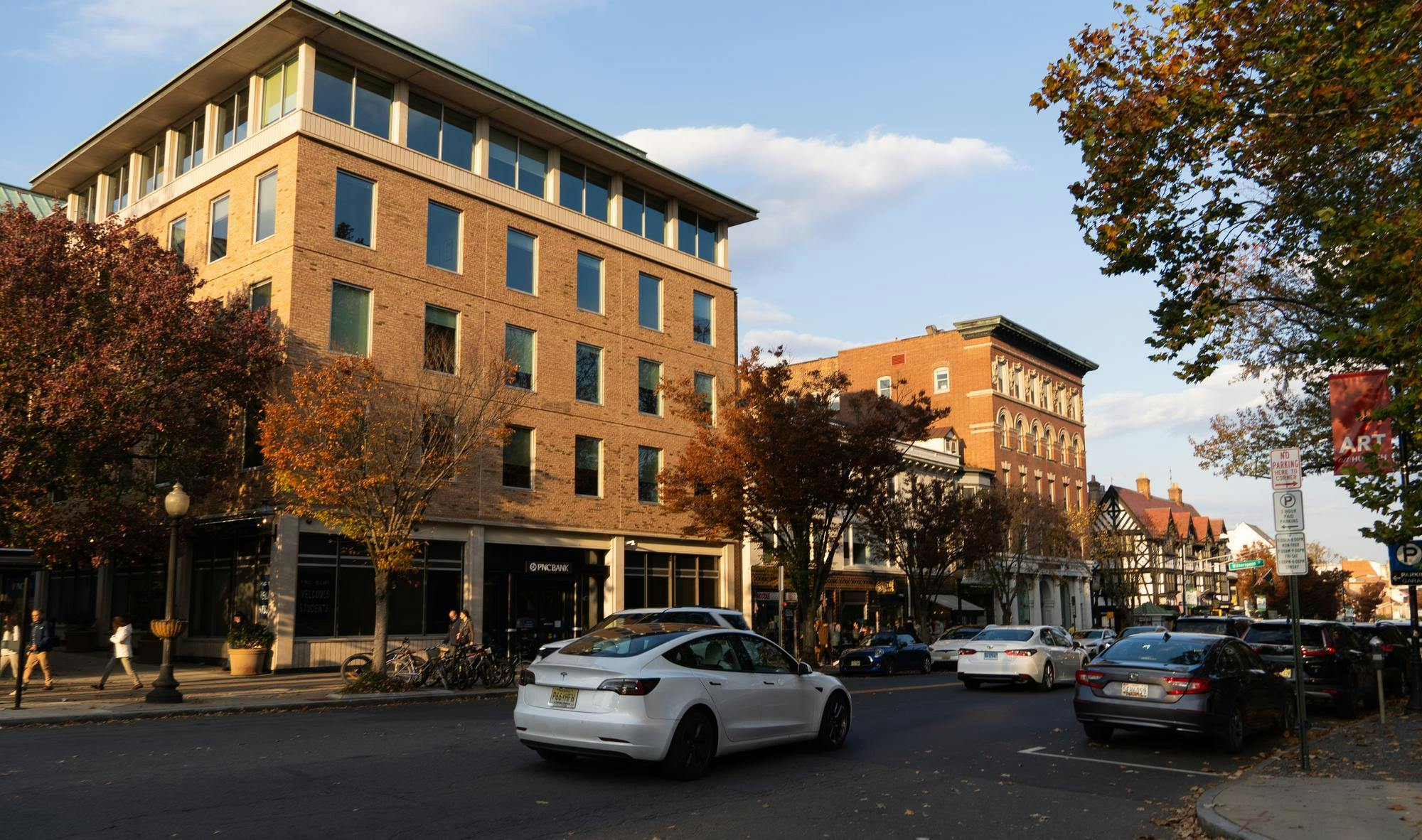 The sun sets over a row of beige buildings on Nassau Street. There is a white Tesla car in the foreground and people are walking along the sidewalk.
