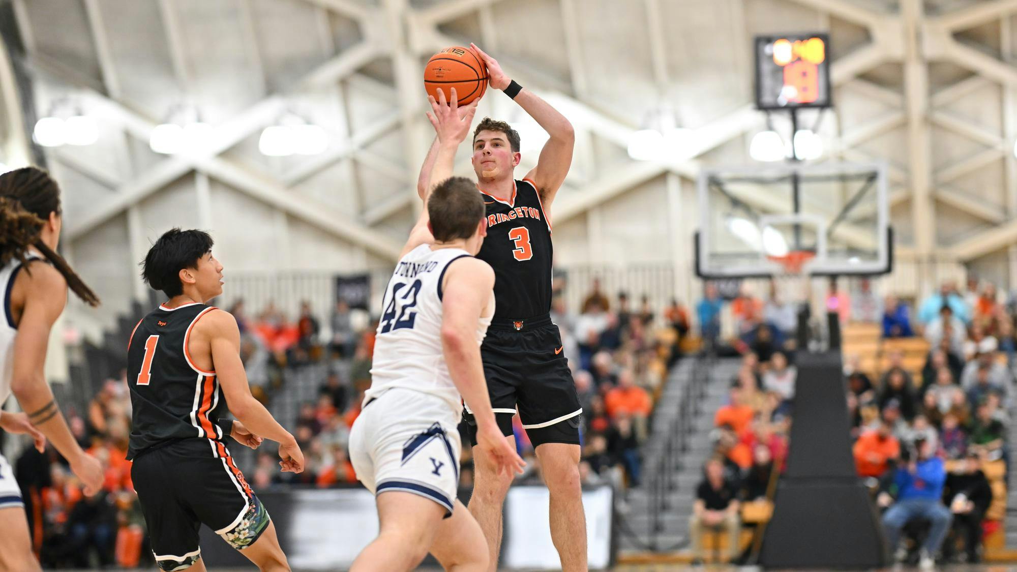 A man shoots a basketball over the outstretched arms of the defender as the crowd watches on. 