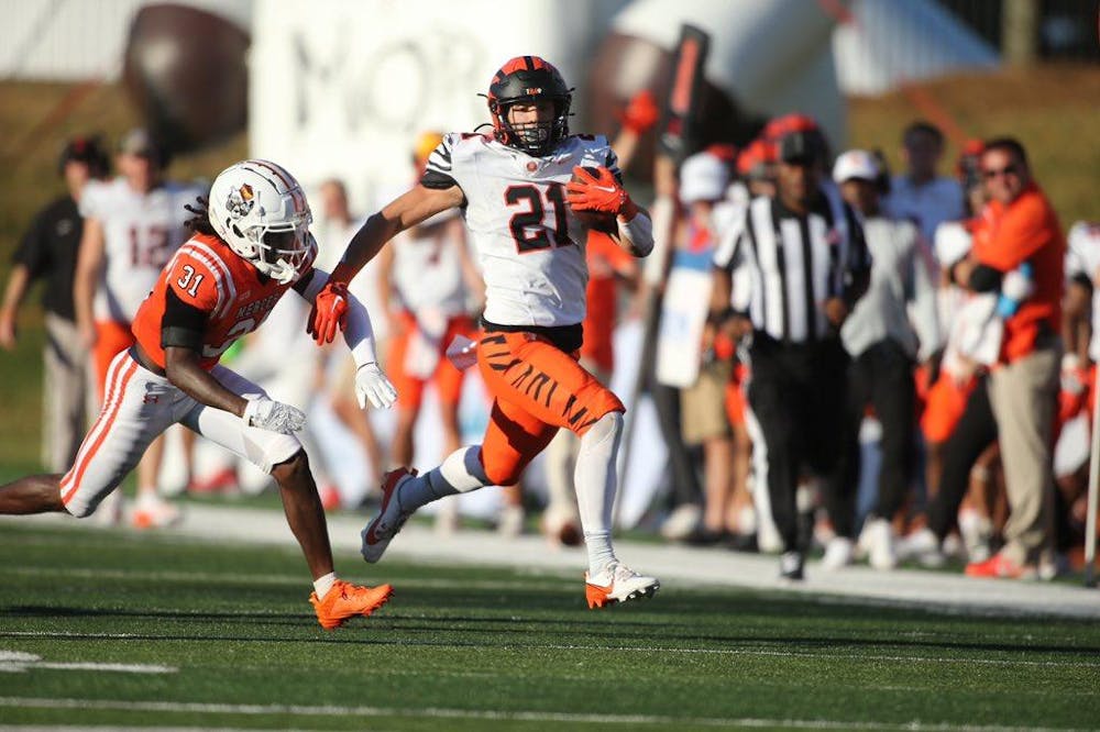 A man wearing an orange and black football jersey with a helmet on running on a field with a football in his hand as he is chased down by a defender. 