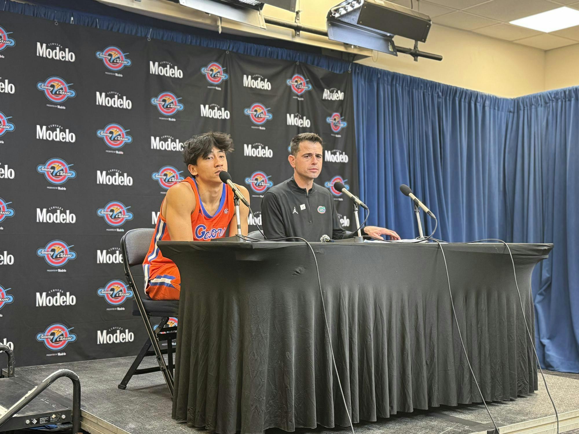 A Florida basketball player and a coach sit at a press conference table with microphones, in front of a Modelo-branded backdrop and blue curtains.