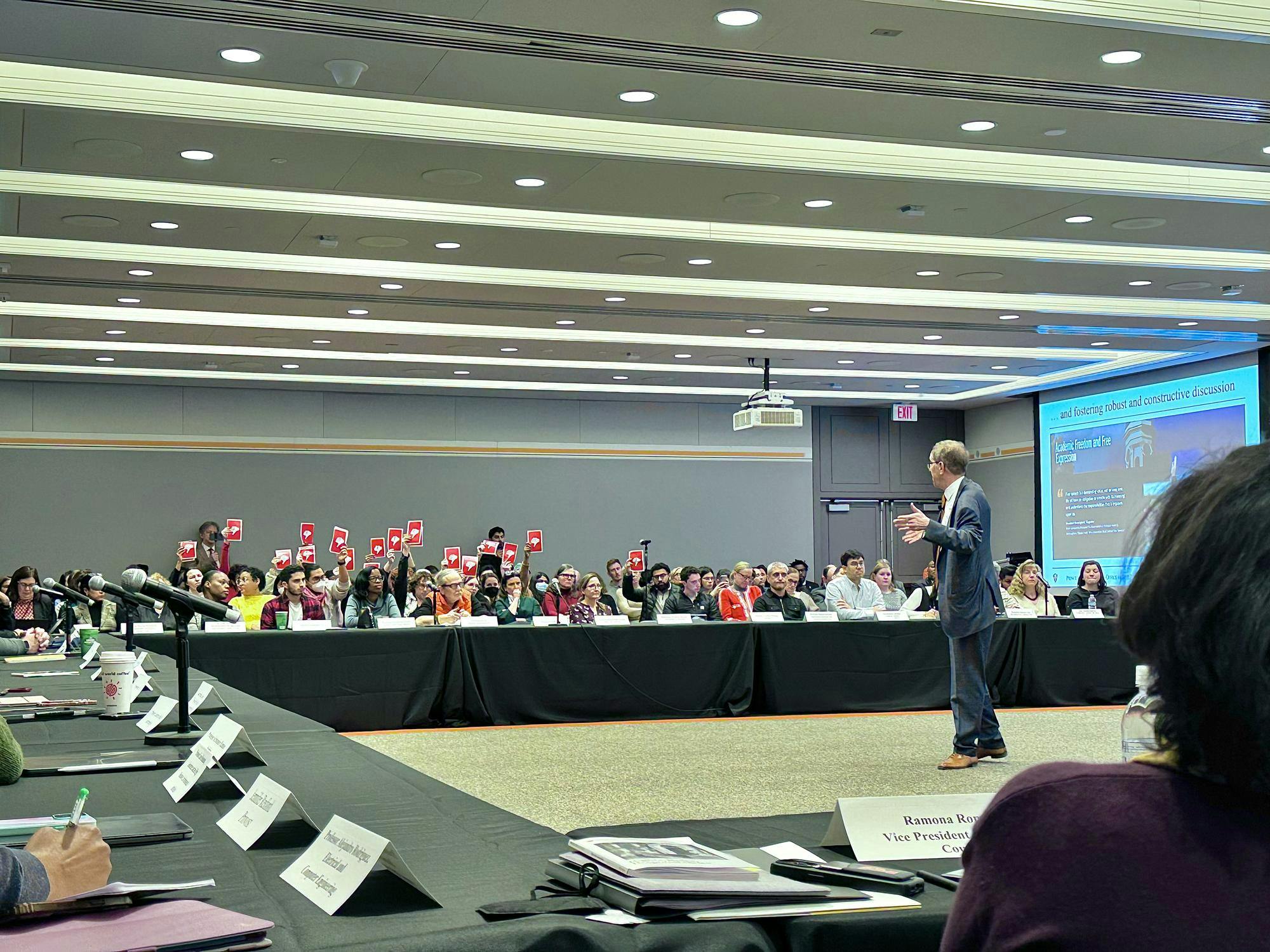 Protestors hold up signs that have red thumbs down on them in the background of an official meeting. A man in a suit speaks to people seated at tables. 