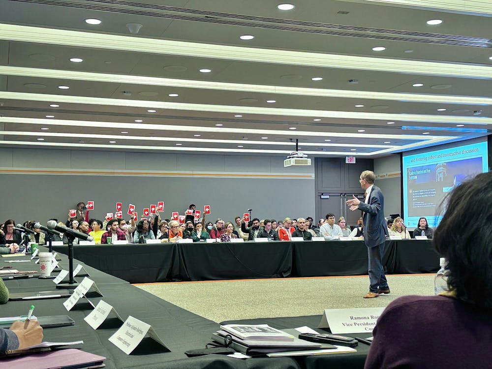 Protestors hold up signs that have red thumbs down on them in the background of an official meeting. A man in a suit speaks to people seated at tables.