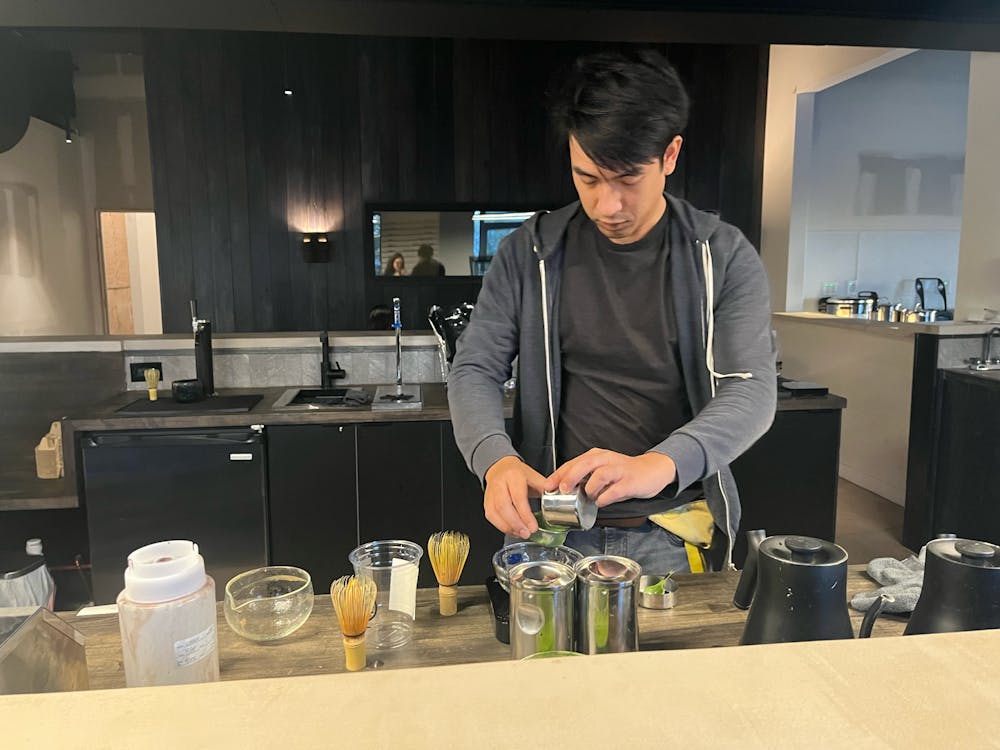 A man is holding two small cups over a bowl as he is mixing ingredients to make a matcha beverage. 