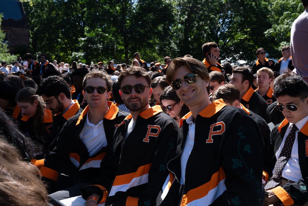 Three students posing on chairs on an outdoor grass lawn wearing sunglasses and an orange and black themed beer jacket. 