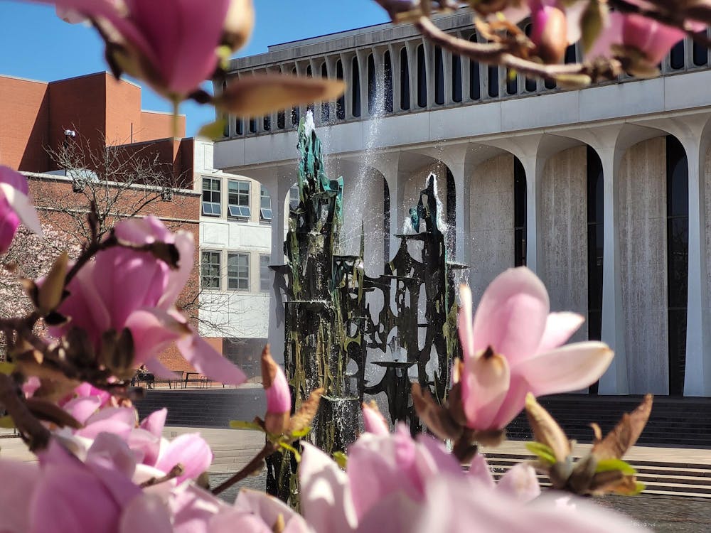 A large fountain and a white building with columns, with pink flowers in the foreground