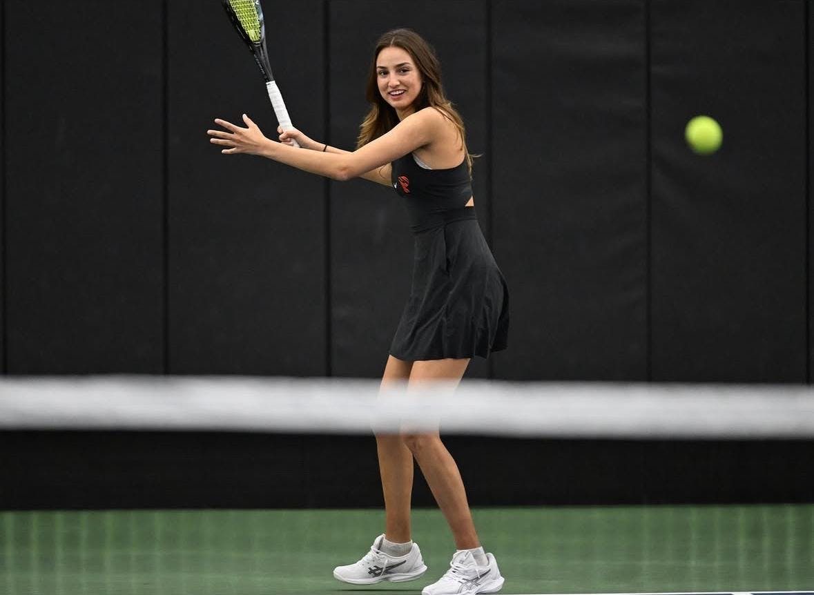 Female tennis player with dark brown hair wearing black uniform and white shoes, smiling while swinging a tennis racket on one side of a green tennis court, black wall in the background