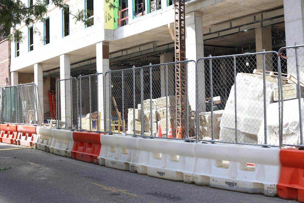 A construction site of a multi-story building with barriers and chain-link fencing surrounding the site. Building materials and equipment are visible inside the fenced area and the building under construction has exposed columns and unfinished walls.