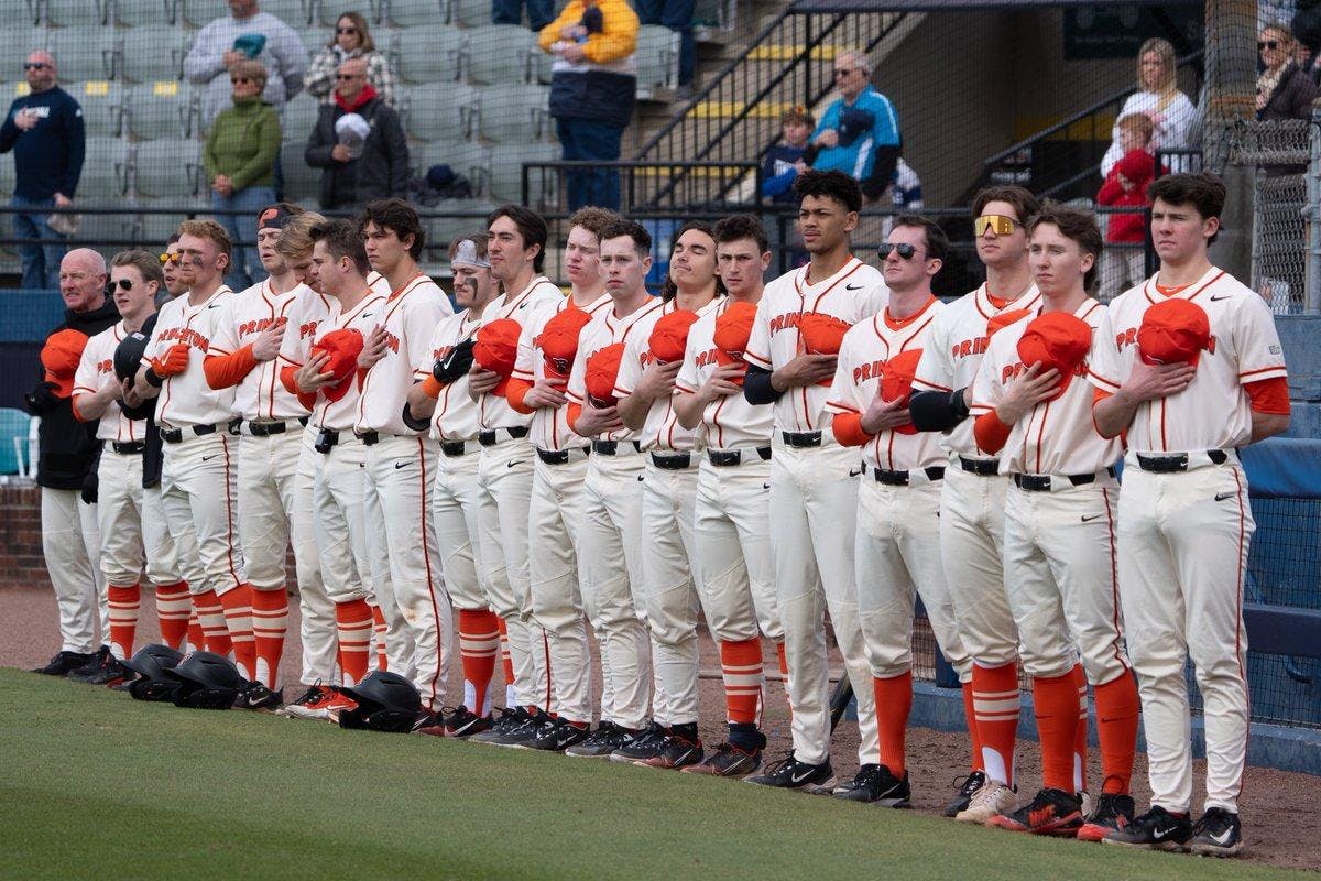 Men in white uniforms line up on the side of a field with hats over hearts for the national anthem.
