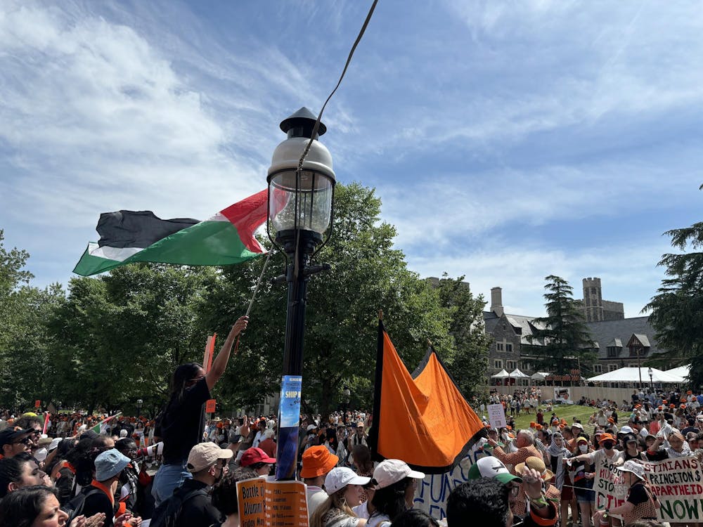 In the foreground, a lamp pole with a Palestinian flag next to it. In the background, a large number of protesters holding signs surrounding a parade route. In the far background, a gray stone building that is Whitman College and a green tree.