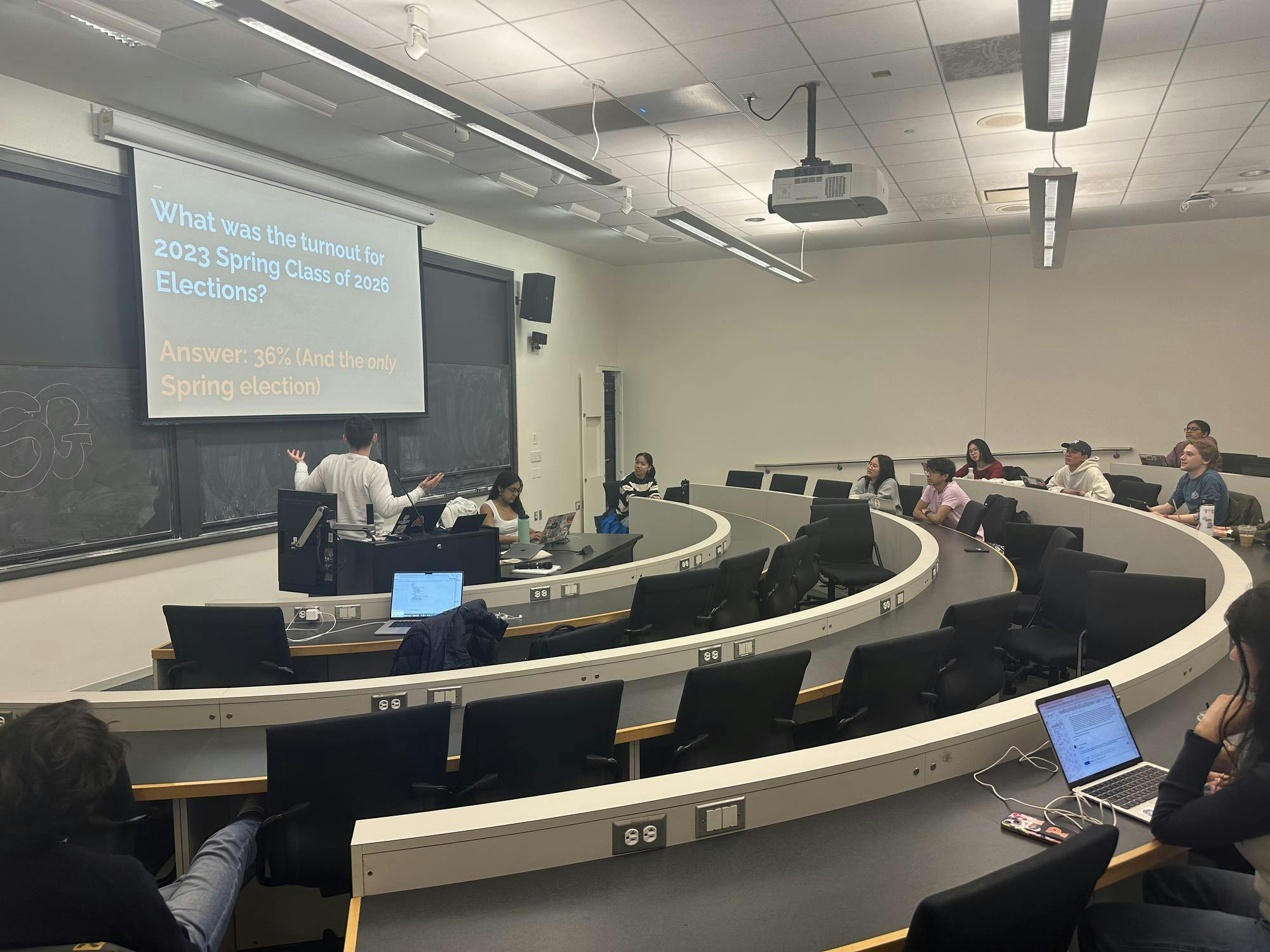 Image of the room with 10 people are in the picture, watching a person give a presentation on election turnout on the left-hand side.