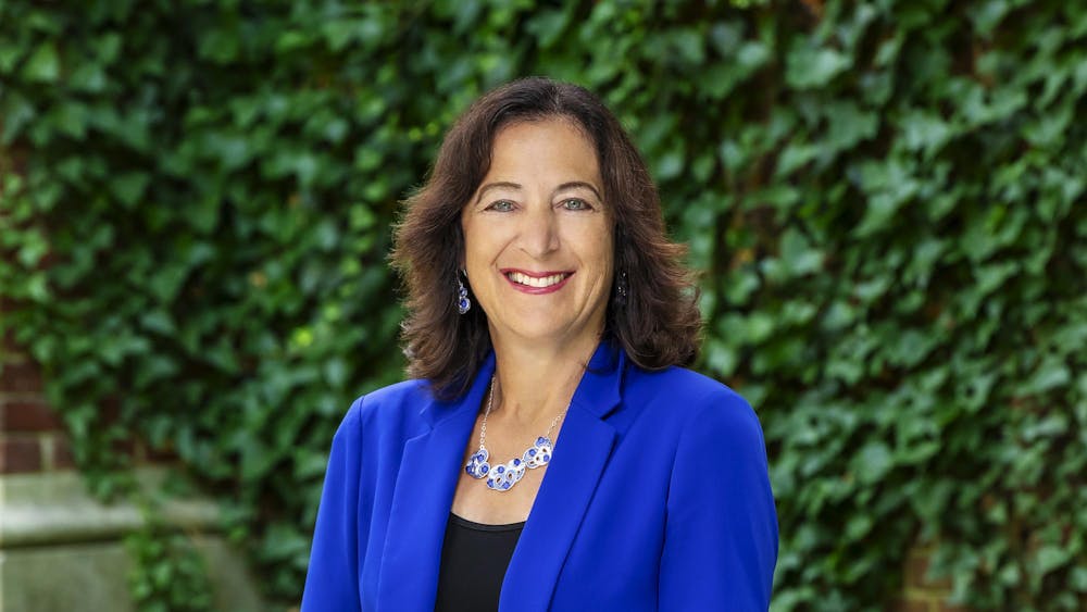 A woman with brown hair wearing a blue blazer and blue and white jewelry, standing in front of ivy-covered walls.