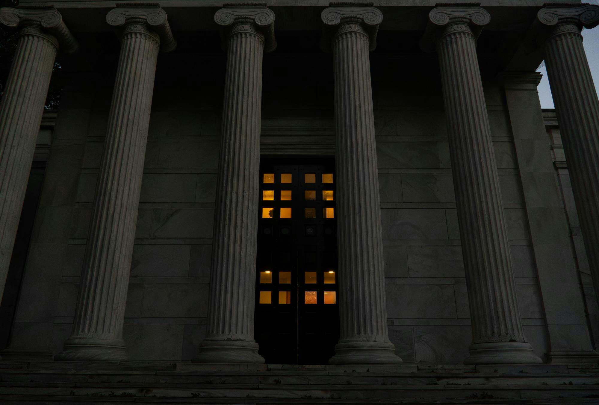A photo of columned building during dusk, with an orange glow behind the door's windows.